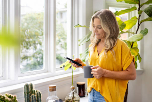 A woman in a yellow shirt holds a smartphone and a cup, standing near a window surrounded by plants.